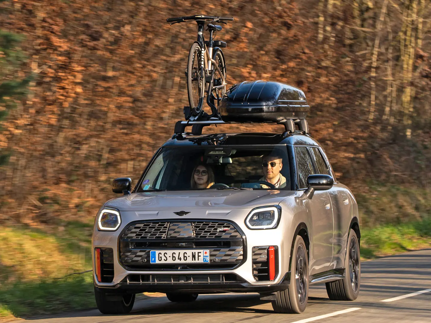Silver fényezésű MINI Countryman Cooper egy hegyi úton, MINI tető-kereszttartóval, MINI tetődobozzal, egy kerékpárral a tetőre szerelt MINI kerékpártartón és MINI John Cooper Works légterelő éllel – elölnézetből. 