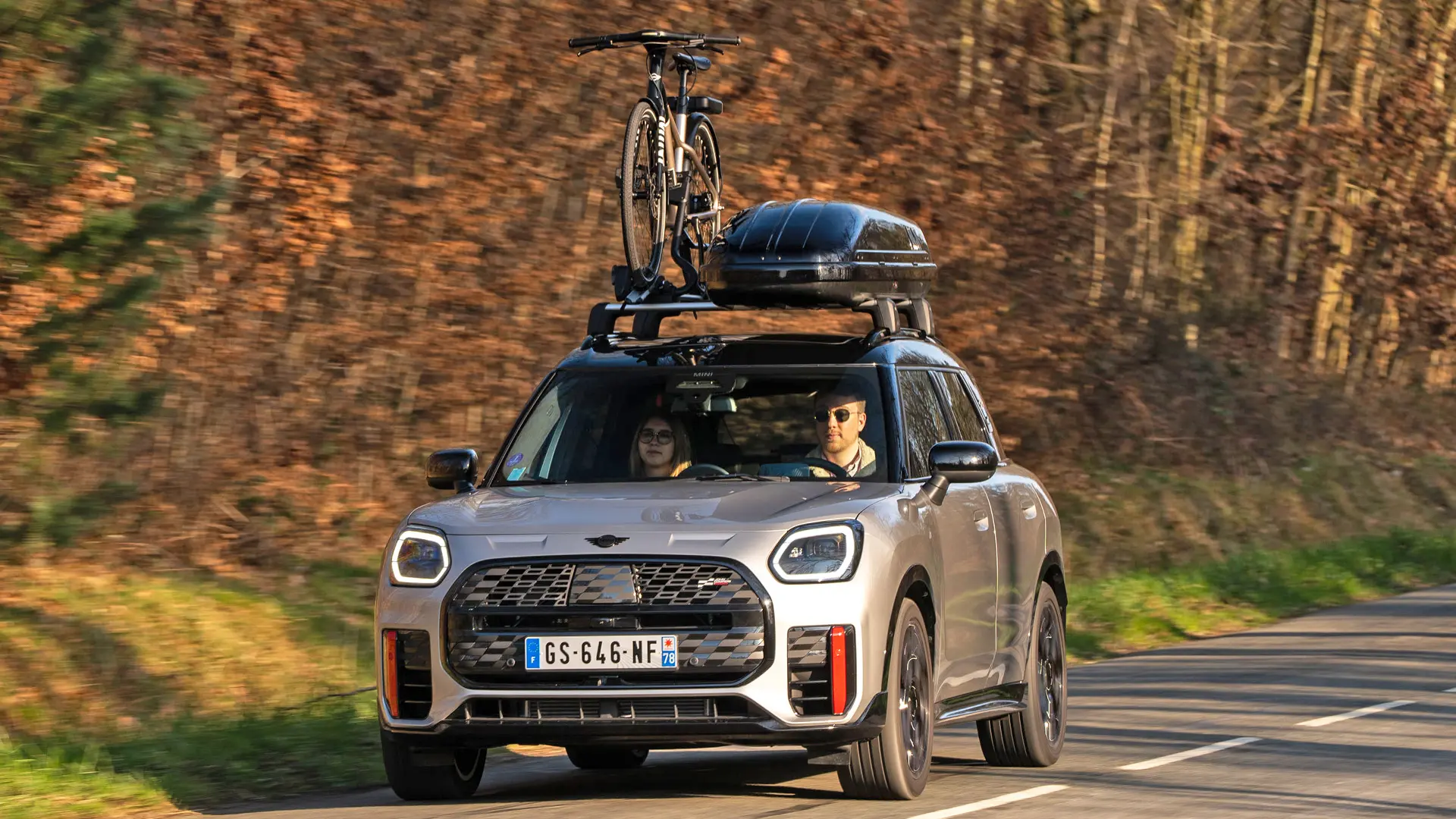 Silver fényezésű MINI Countryman Cooper egy hegyi úton, MINI tető-kereszttartóval, MINI tetődobozzal, egy kerékpárral a tetőre szerelt MINI kerékpártartón és MINI John Cooper Works légterelő éllel – elölnézetből. 