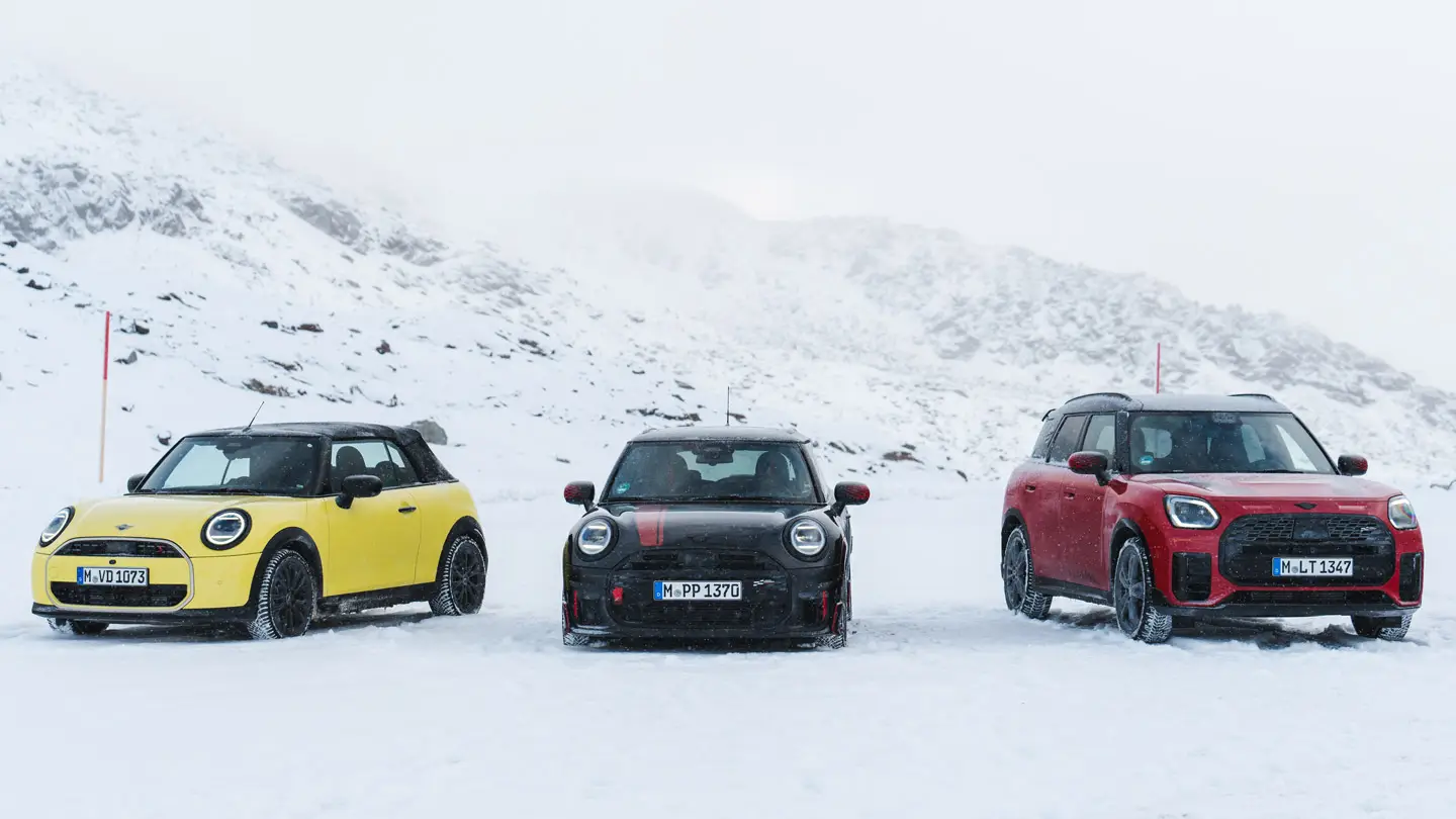 A MINI Cooper Cabrio Sunny Side Yellow, MINI John Cooper Works Midnight Black II és MINI Countryman Cooper Chilli Red fényezéssel – az autók egy havas hegyi úton állnak – elölnézet.
