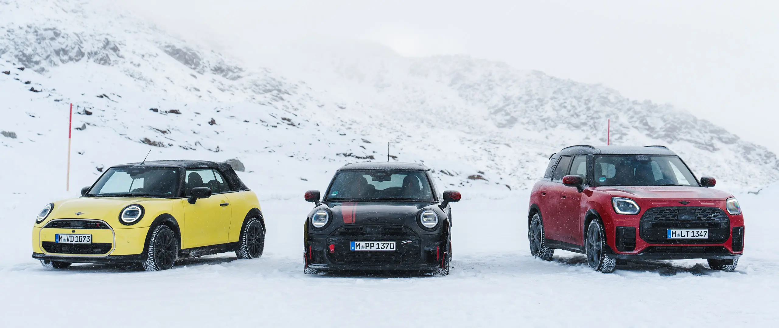 A MINI Cooper Cabrio Sunny Side Yellow, MINI John Cooper Works Midnight Black II és MINI Countryman Cooper Chilli Red fényezéssel – az autók egy havas hegyi úton állnak – elölnézet.