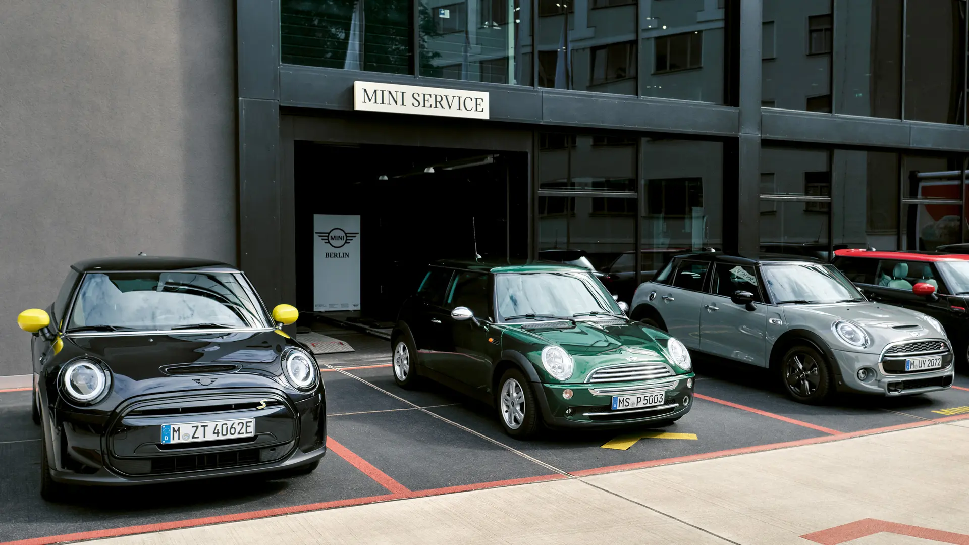 Egy fekete elektromos MINI Cooper S egy MINI márkakereskedés előtt, egy zöld, 3 ajtós MINI Cooperrel, egy ezüstszínű 5 ajtós MINI-vel és egy fekete MINI JCW-vel, szemből. 