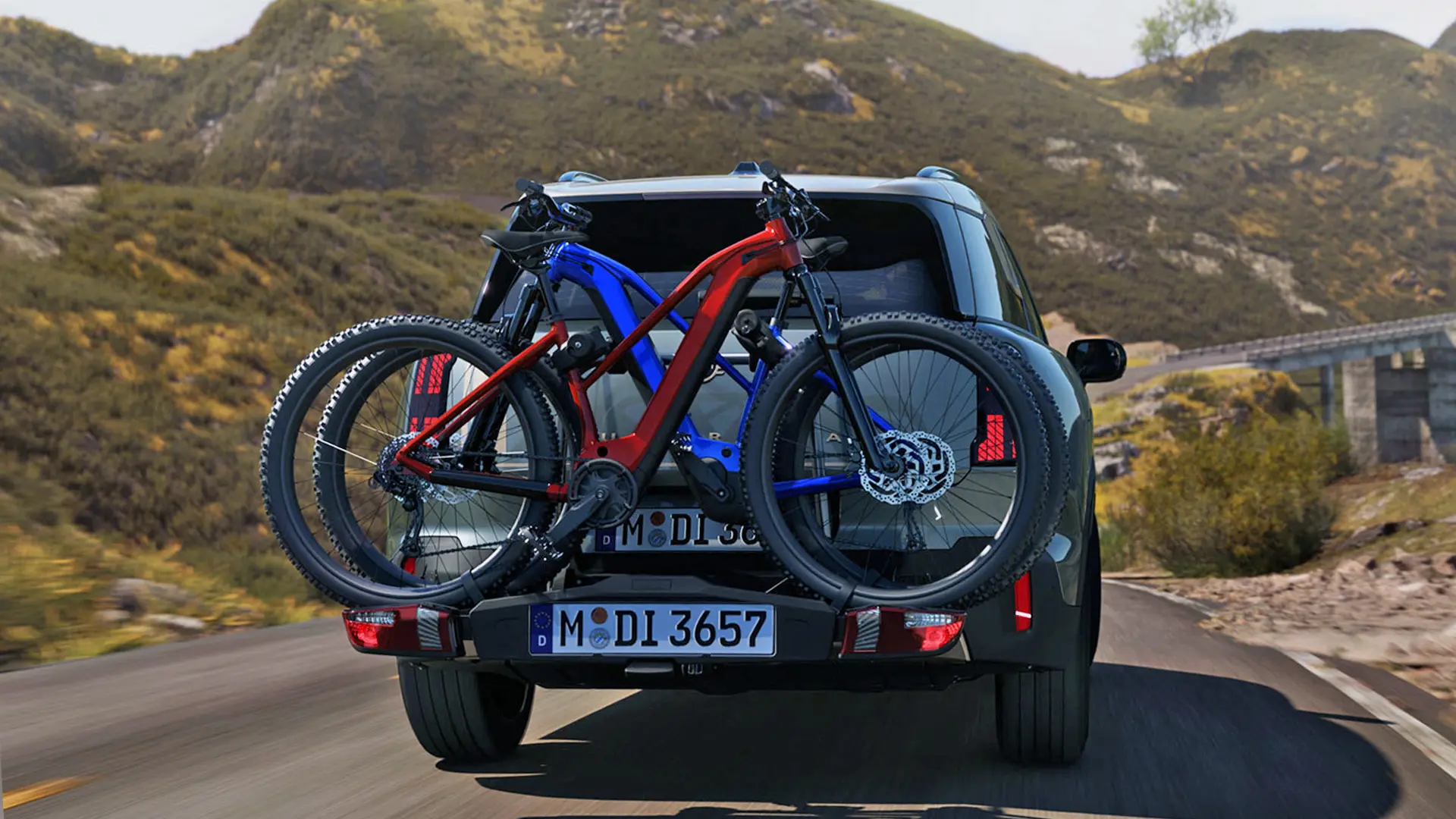 Egy Smokey Green fényezésű, hátsó kerékpártartó-rendszerrel – amelyen két mountain bike van elhelyezve – ellátott, hegyi úton haladó MINI Countryman hátulról.